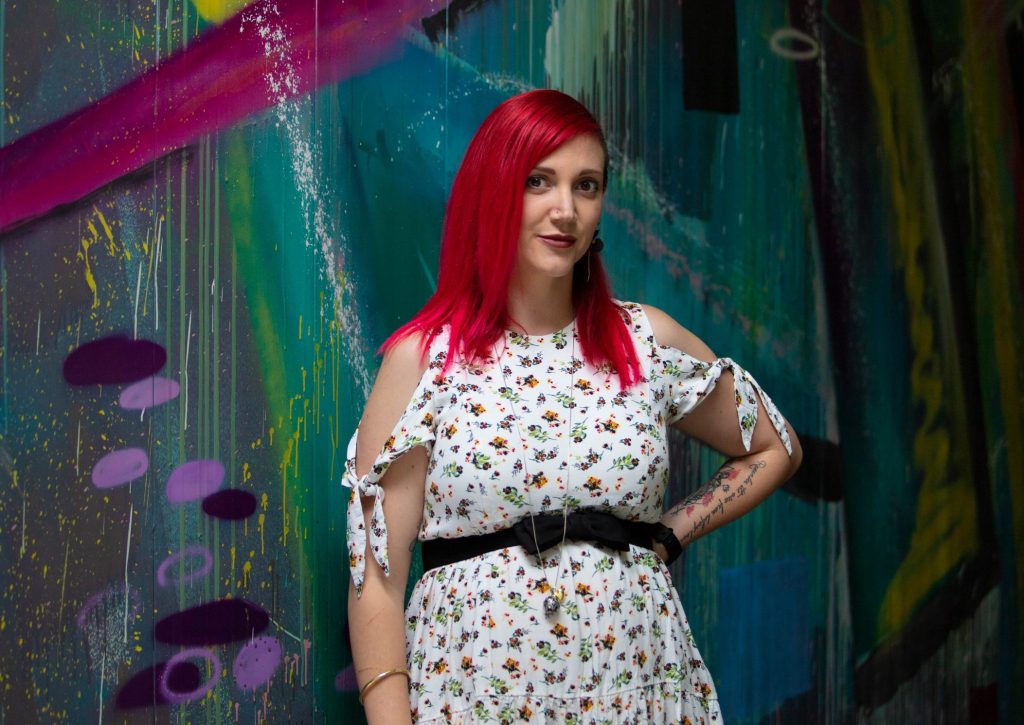 Photo of a woman with bright red hair standing before a colourful wall
